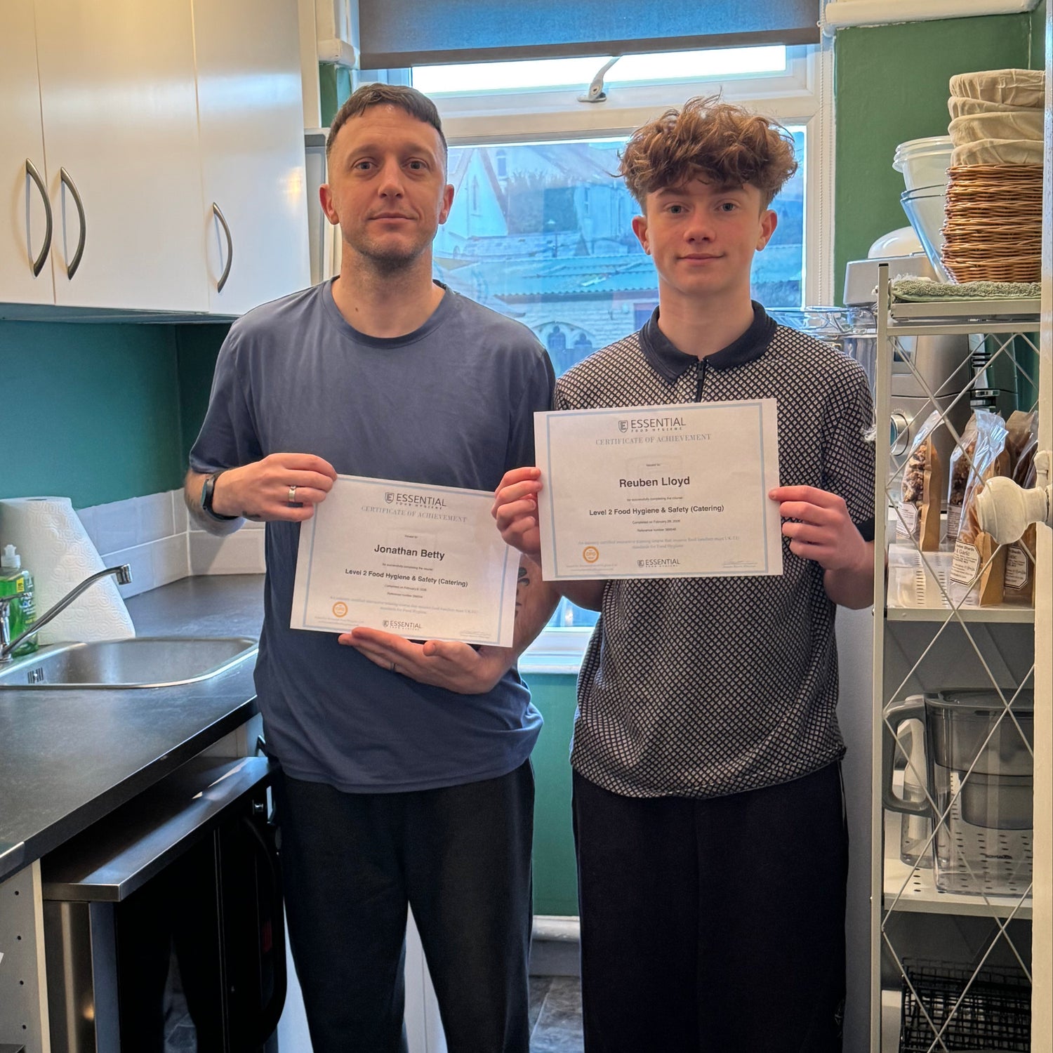 Two people holding certificates in a kitchen setting